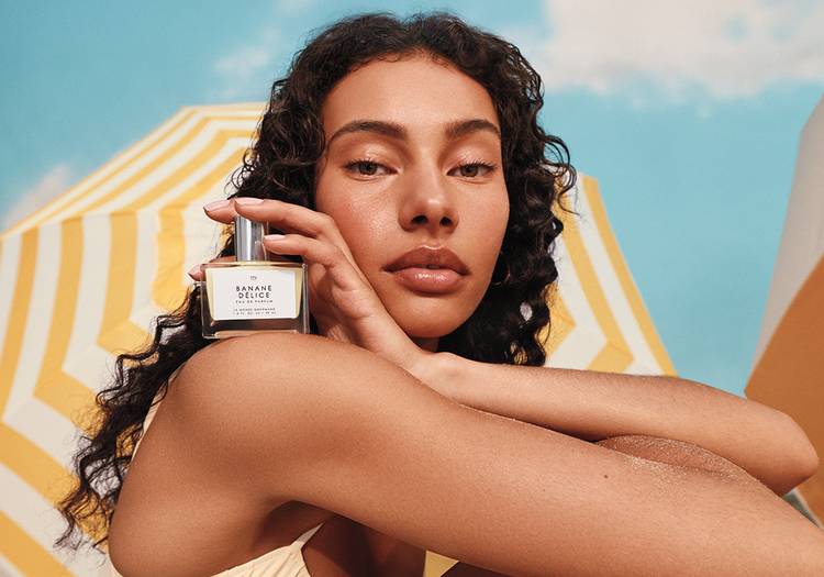 A model holds the Le Monde Gourmand Banane Délice Eau de Parfum against her cheek in front of a yellow-striped beach umbrella.A model holds the Le Monde Gourmand Banane Délice Eau de Parfum against her cheek in front of a yellow-striped beach umbrella.