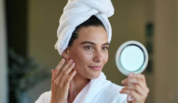Woman in a white robe and hair towel touching her cheek while examining her skin in a handheld mirror