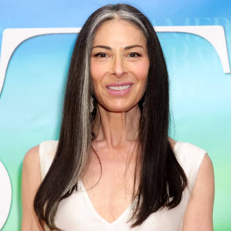 Stacy London smiling at a red carpet event, wearing a white sleeveless top with her dark hair, streaked with gray, worn long and parted down the middle.