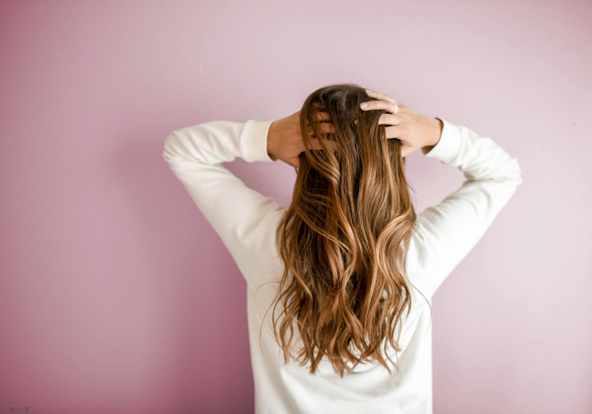 Woman with long, wavy brown and caramel balayage hair seen from behind, running her fingers through her scalp against a mauve background.