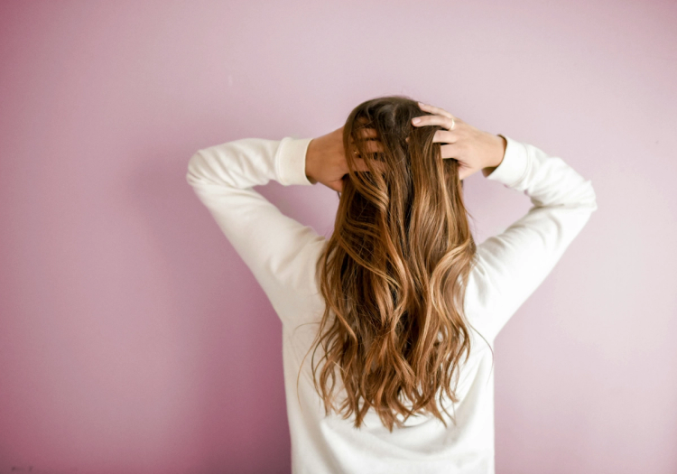 Woman with long, wavy brown and caramel balayage hair seen from behind, running her fingers through her scalp against a mauve background.