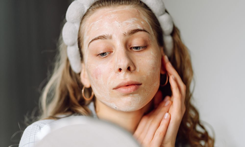 A portrait of a young woman looking at her skin covered with a white mask in a small mirror while sitting in a cozy room. Beauty, facial care, cosmetology concept.
