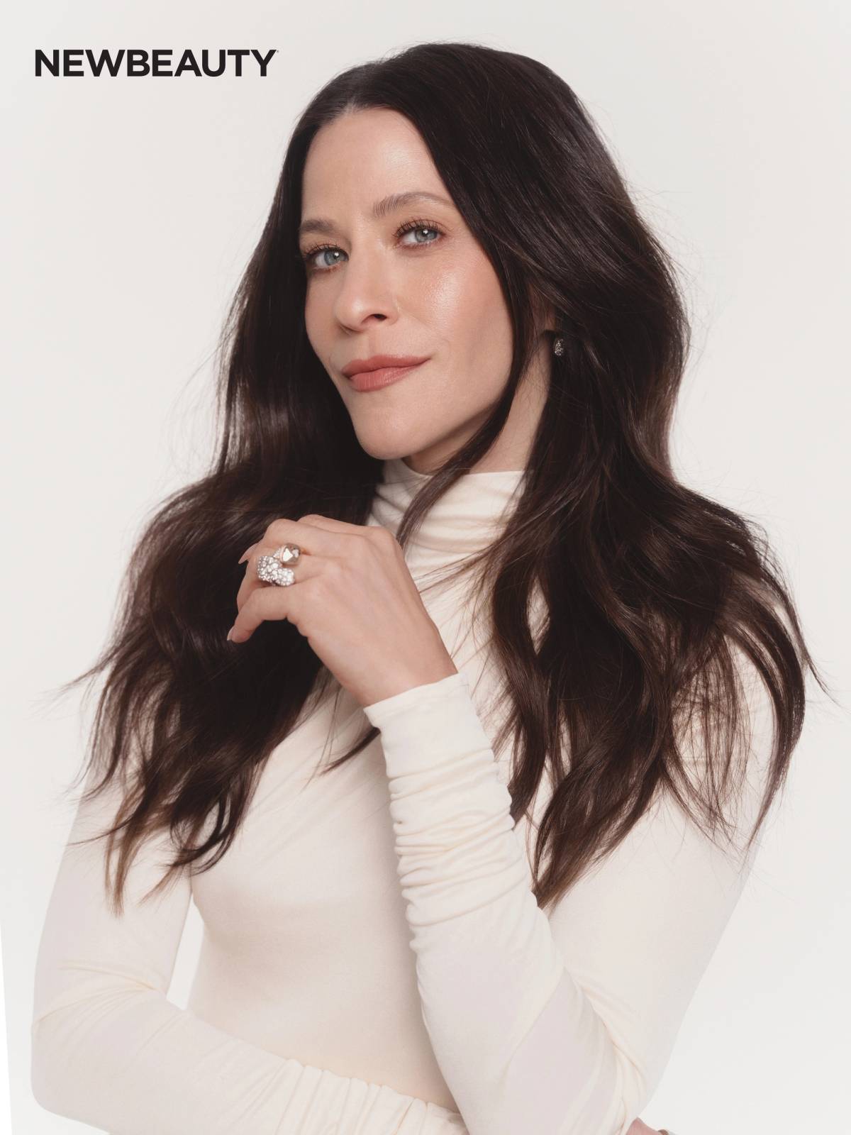 Jackie Tohn in a cream turtleneck, wearing a sparkling statement ring and smiling softly at the camera against a white background.