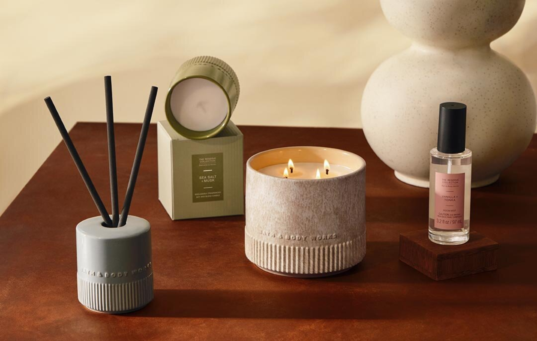 Bath & Body Works Reserve Collection products styled on a wooden table, including a lit three-wick candle, a reed diffuser, a boxed Sea Salt + Musk candle and a Vanilla + Tonka room mist.