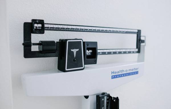 Close-up of a physician’s balance-beam scale against a white wall.