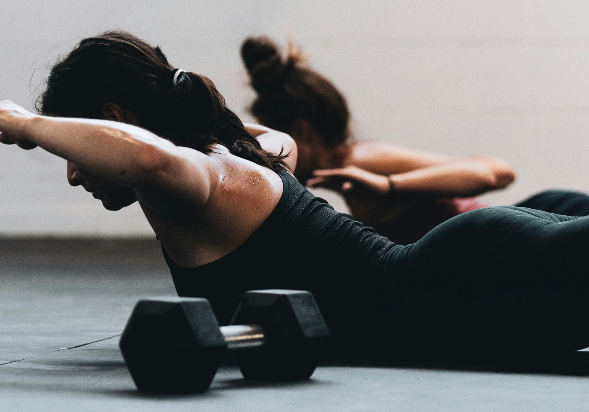Two women doing a strength-training workout on mats with a dumbbell in the foreground.