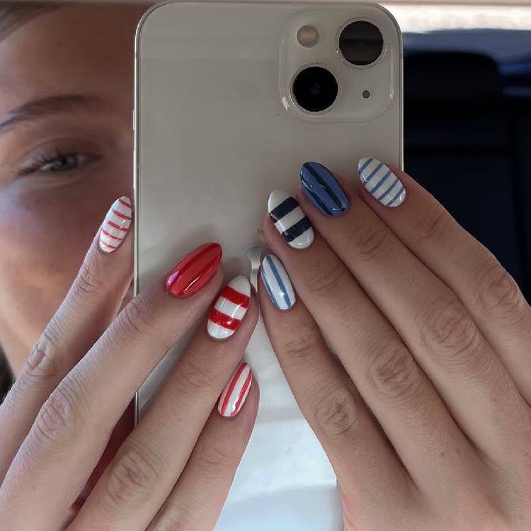 A person holding a phone up to a car mirror showing red and white striped nails on one hand and navy and white striped nails on the other, in an alternating mixed stripe manicure