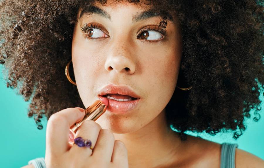 Model applying a rose-toned lipstick in a gold tube, shown against a teal studio backdrop