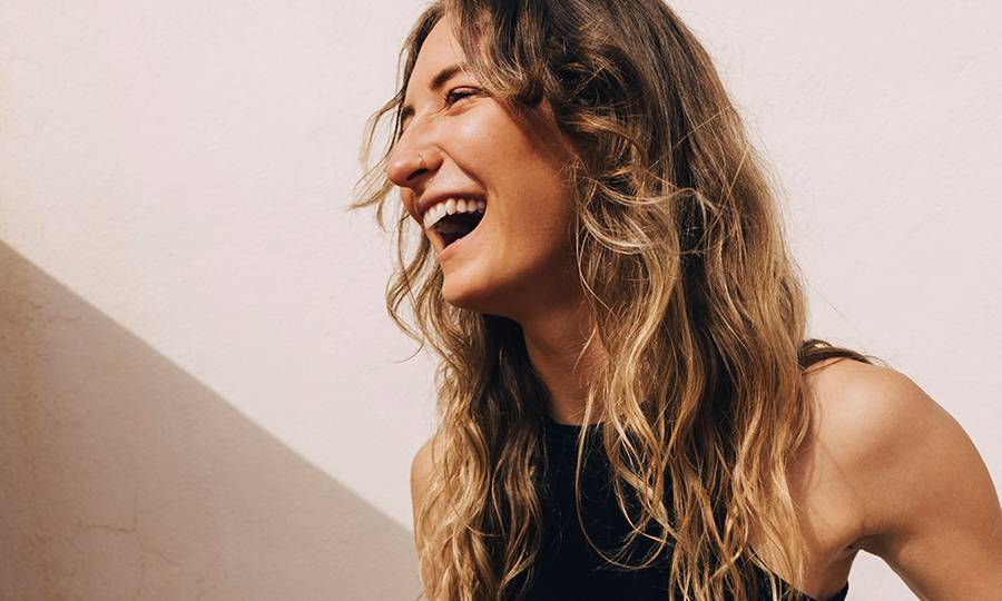Young woman laughing while holding yoga mat and water bottle near wall