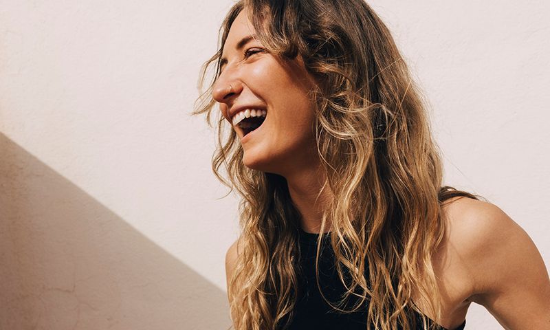 Young woman laughing while holding yoga mat and water bottle near wall