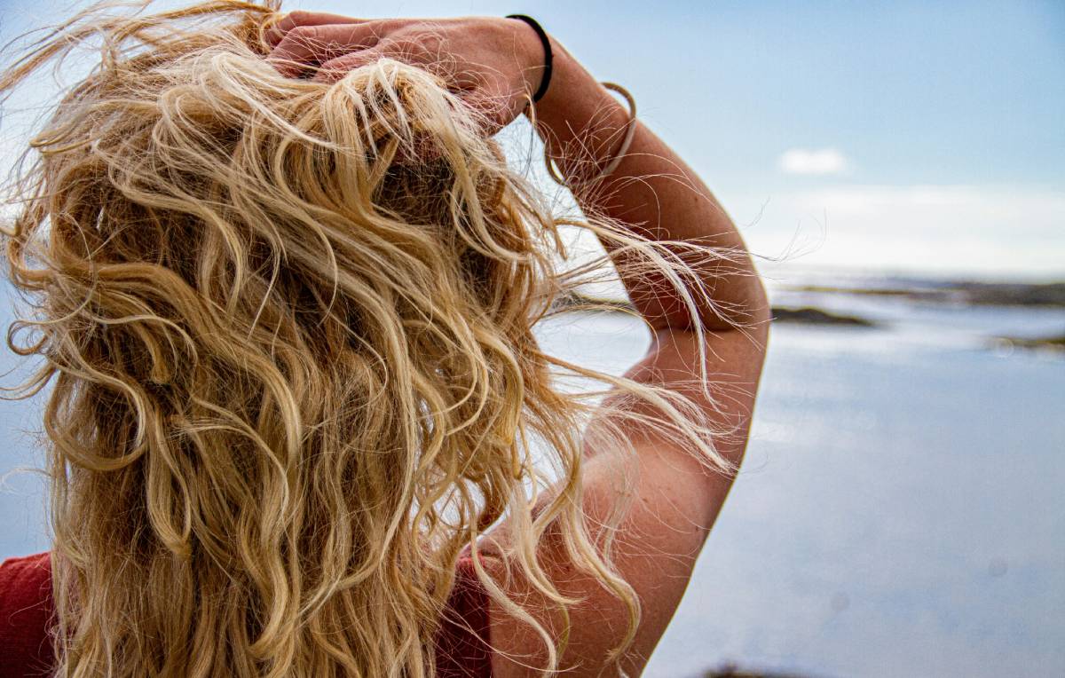 Woman with tousled, wavy blonde hair running her fingers through it against a backdrop of the ocean and sky.