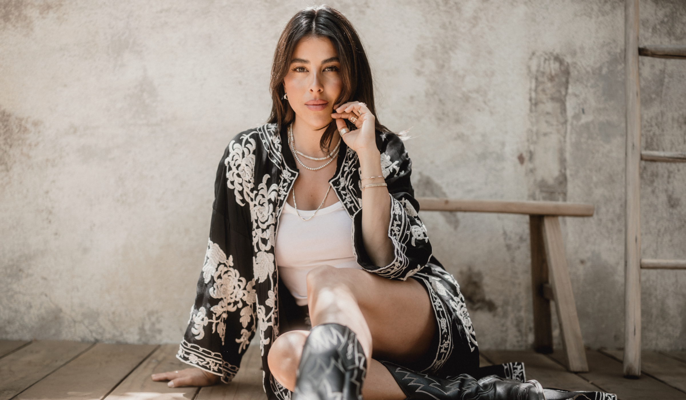 Daniella Monet seated on a wood floor in an embroidered black kimono jacket, white tank and black cowboy boots, hand at her cheek.