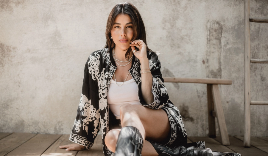 Daniella Monet seated on a wood floor in an embroidered black kimono jacket, white tank and black cowboy boots, hand at her cheek.