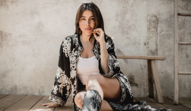 Daniella Monet seated on a wood floor in an embroidered black kimono jacket, white tank and black cowboy boots, hand at her cheek.