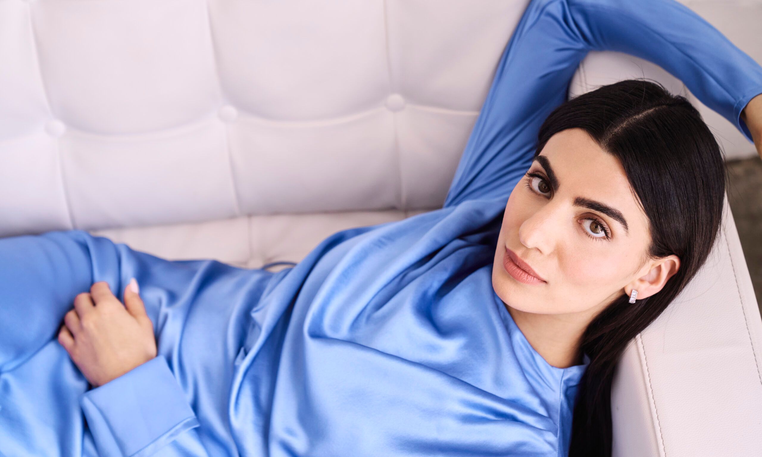 Claudia Doumit lounging on a sofa in a blue long-sleeved silk dress