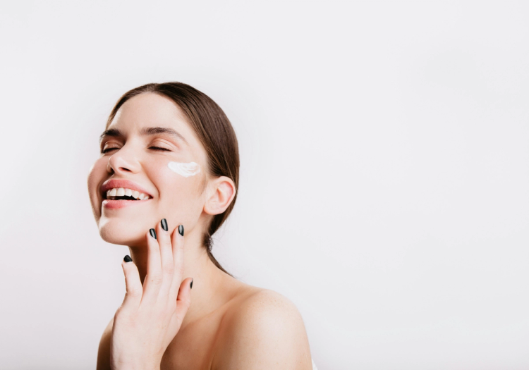 Smiling woman with eyes closed and a swipe of white cream on her cheek, touching her jaw against a white background