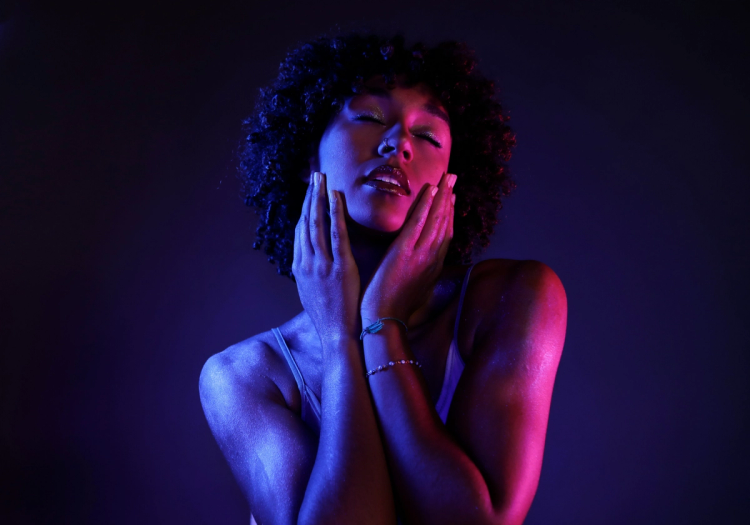 Close-up of a woman with curly hair, eyes closed, holding both hands to her cheeks under blue and pink lighting.