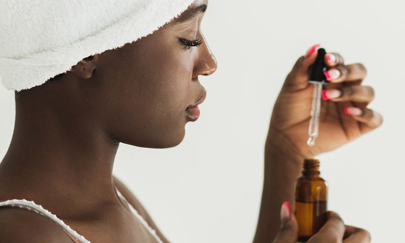 Happy young black woman with covered white towel on head applying serum on face with closed eyes. Beautiful afro-american woman apply serum vitamin C, argan oil or tea tree ingredients in pippette dropper under eye, treating skin for nourishing and moisturizing effect against white background. Beauty and skin care concept.