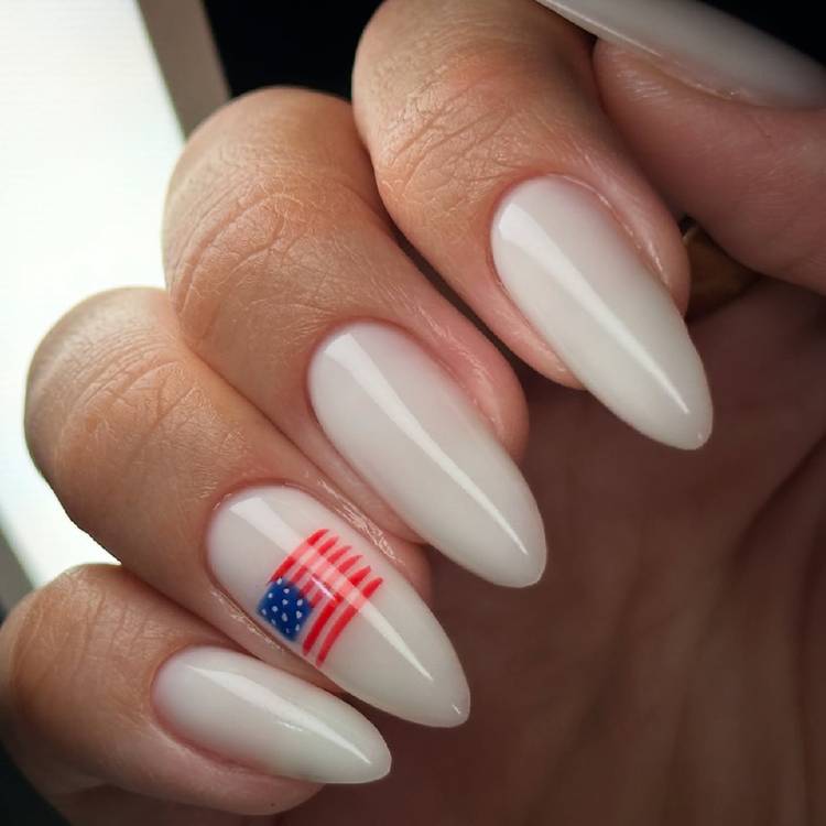 Close-up of almond-shaped milky white nails with a small American flag nail art accent on the ring finger