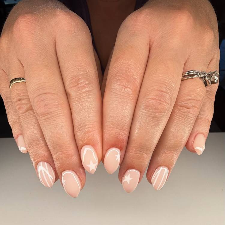 Close-up of two hands resting on a white surface with soft nude almond nails featuring white star accents and fluid swirl nail art, with gold and silver rings visible on each hand