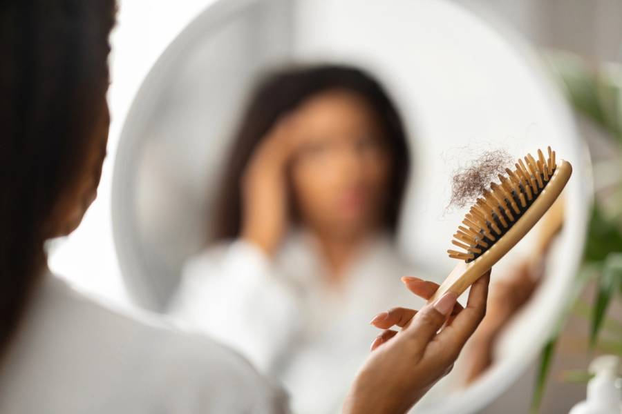 Woman holding a wooden hairbrush filled with loose hair while looking at her reflection in a bathroom mirror.