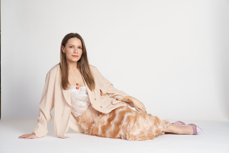 Bethany Joy Lenz posed on a studio floor in a neutral-toned outfit, reclining with a relaxed, confident expression for a portrait accompanying her NewBeauty interview.