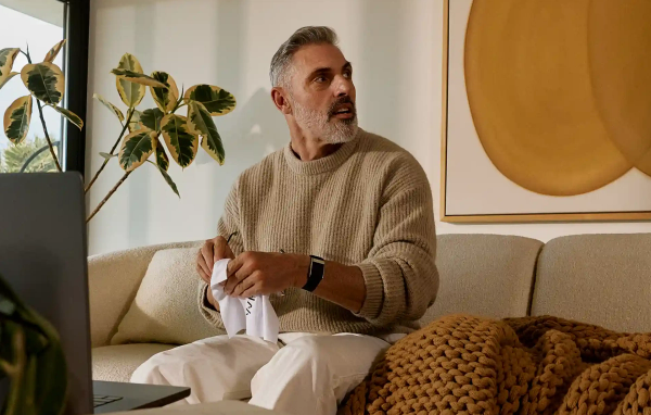 A gray-haired, bearded man in a beige sweater sits on a couch wearing a WHOOP wearable on his wrist