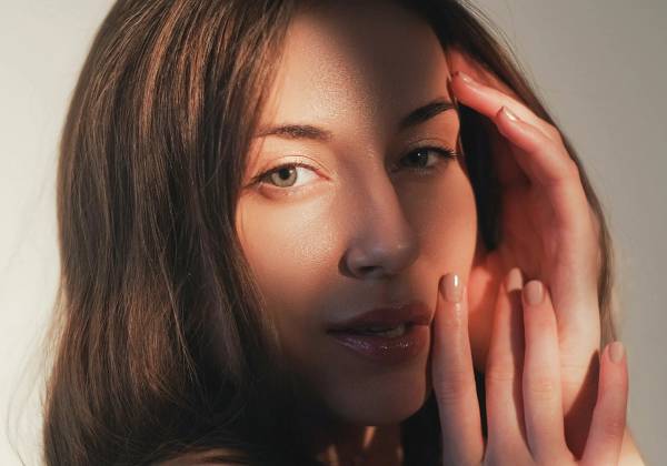 12:05 PM A woman with glowing, even-toned skin holds her hand to her face in warm light