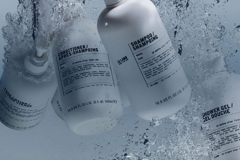 Le Labo Basil shampoo, conditioner and lotion bottles surrounded by splashing water, highlighting the brand’s minimalist packaging and fresh locker-room aesthetic.