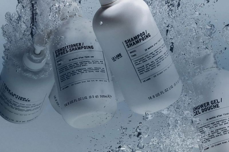 Le Labo Basil shampoo, conditioner and lotion bottles surrounded by splashing water, highlighting the brand’s minimalist packaging and fresh locker-room aesthetic.