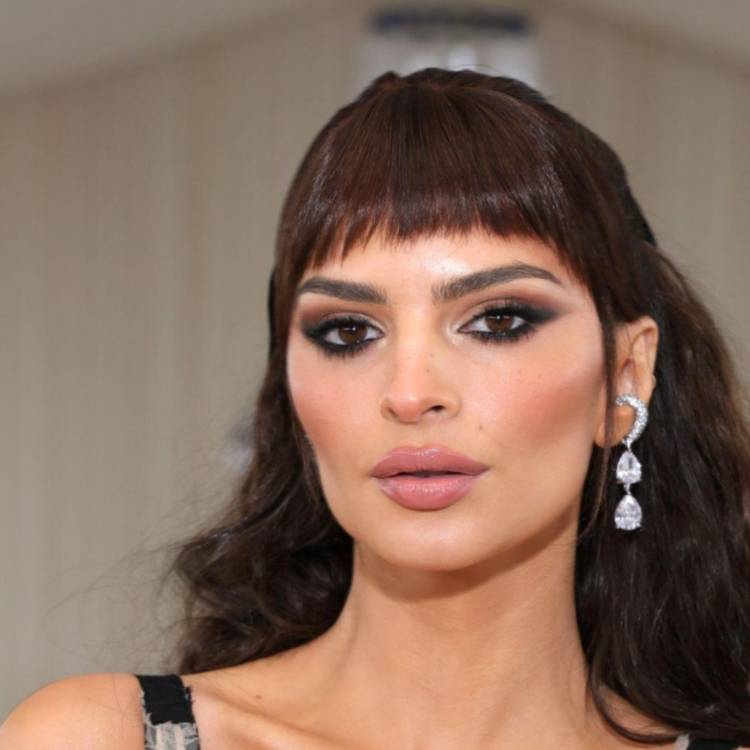 Emily Ratajkowski in black two-piece outfit poses in front of a pink background