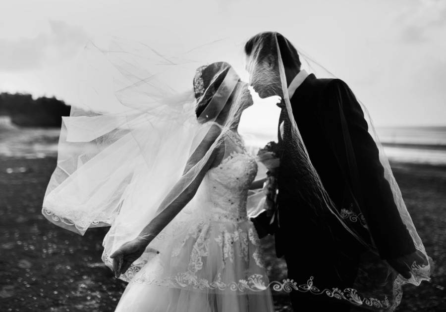 Bride and groom kissing on a beach, her veil blowing dramatically in the wind