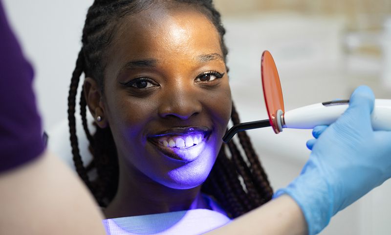 Portrait of cheerful black woman, radiant with bright smile, looking at camera while sitting in dentist chair in clinic and receiving high-quality filling procedure with use of ultraviolet lamp. White male dentist, wearing gloves, holding tool and carefully guiding light, showcasing advancements in modern dentistry.