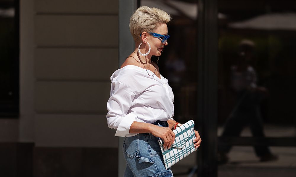 Sophisticated woman with short hair in white shirt and cargo jeans pants, fashionable accessories earrings, stylish glasses and clutch walking outdoors in city. Close up Fashion style.