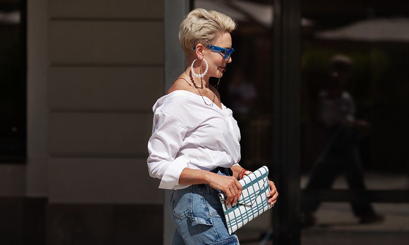 Sophisticated woman with short hair in white shirt and cargo jeans pants, fashionable accessories earrings, stylish glasses and clutch walking outdoors in city. Close up Fashion style.