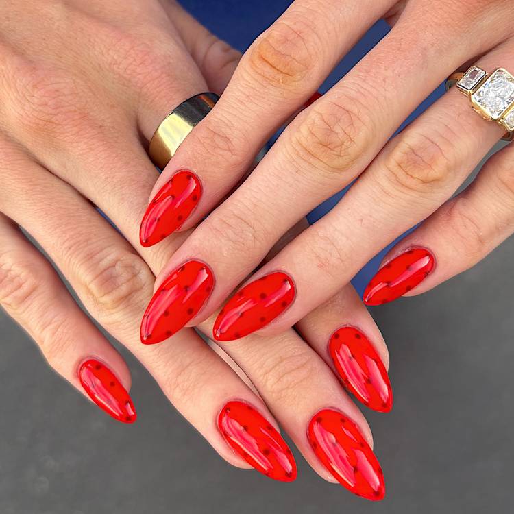 Close-up of two hands with glossy bright red stiletto nails featuring scattered dark red micro dot nail art, with gold and diamond rings visible