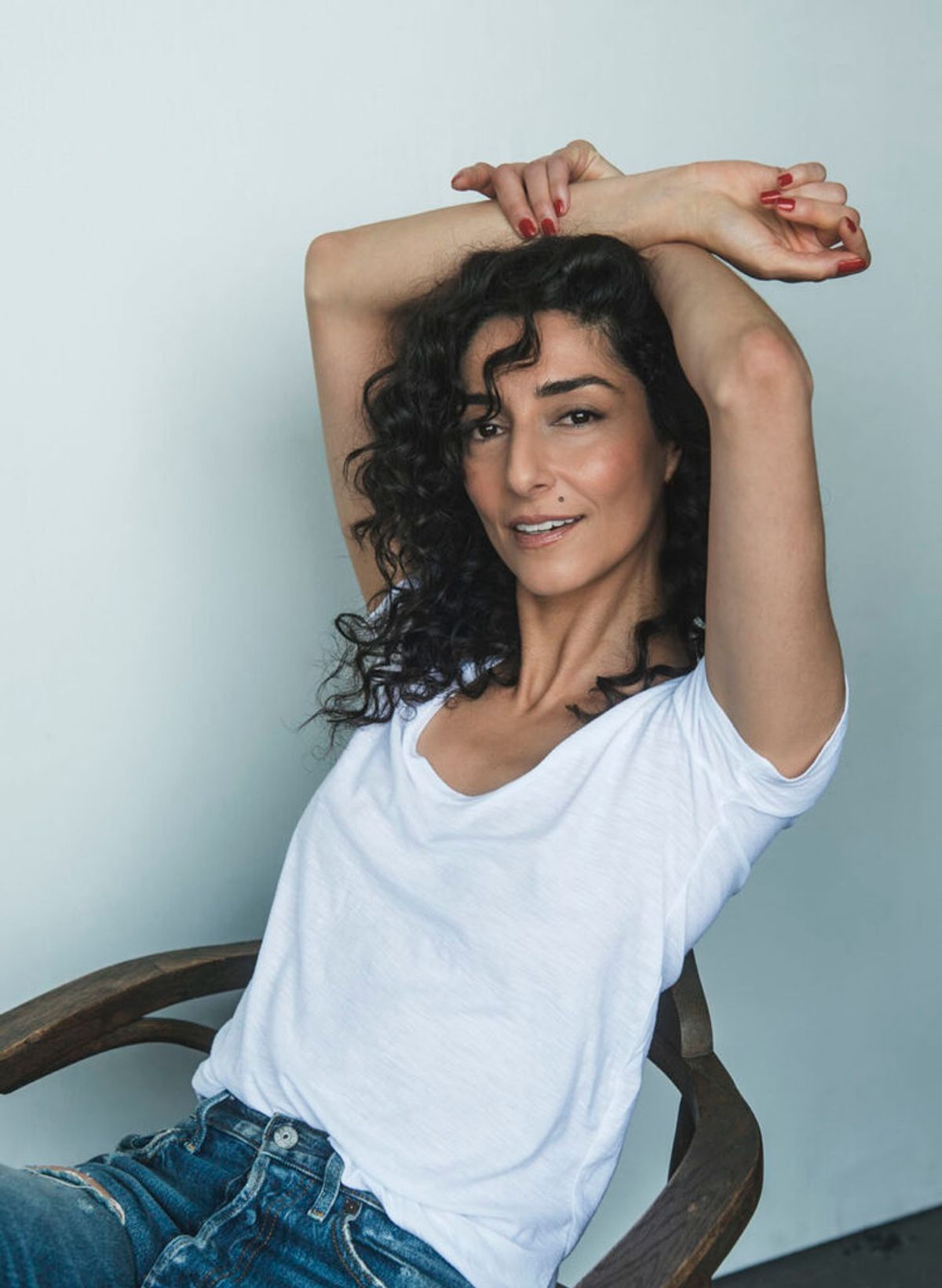 Necar Zadegan has curly hair and is sitting with her hands over her head wearing a white t-shirt and jeans.