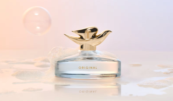 Dove Original Fragrance bottle with a gold dove-shaped cap, resting in soap suds on a wet, glossy surface beside a floating bubble against a soft peach-and-lavender backdrop.