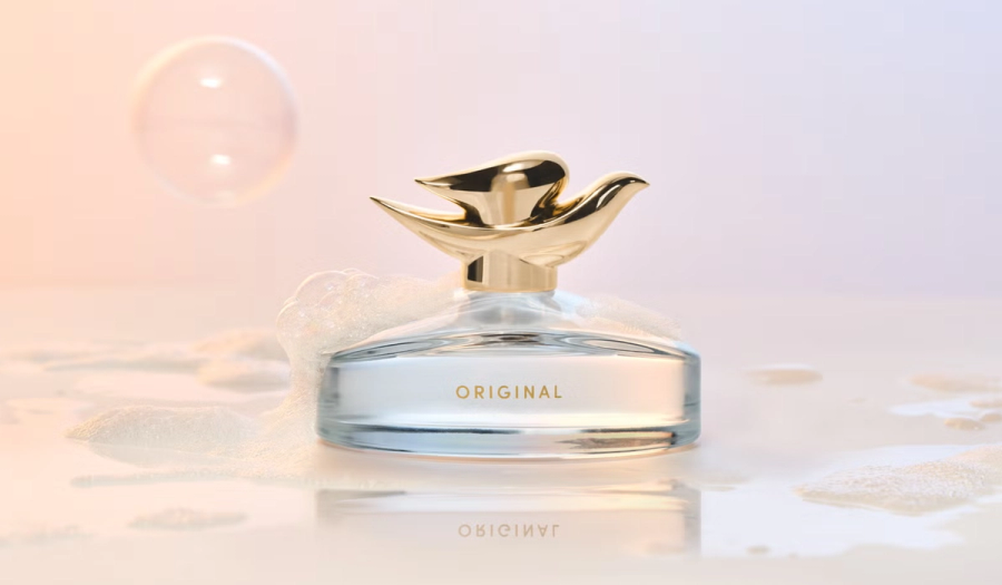 Dove Original Fragrance bottle with a gold dove-shaped cap, resting in soap suds on a wet, glossy surface beside a floating bubble against a soft peach-and-lavender backdrop.