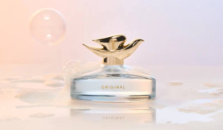 Dove Original Fragrance bottle with a gold dove-shaped cap, resting in soap suds on a wet, glossy surface beside a floating bubble against a soft peach-and-lavender backdrop.