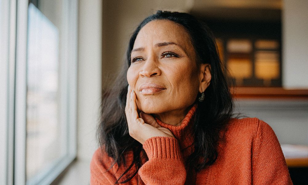 A woman in a cozy orange sweater gazes thoughtfully out of a window, resting her chin on her hand. Soft lighting highlights her serene expression and the warm tones of the room around her.
