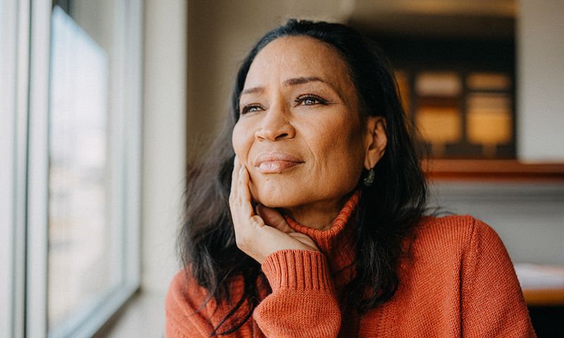 A woman in a cozy orange sweater gazes thoughtfully out of a window, resting her chin on her hand. Soft lighting highlights her serene expression and the warm tones of the room around her.