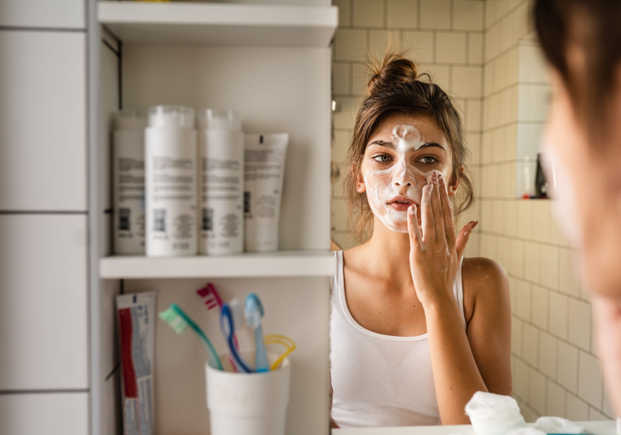 Young woman washing her face with a foaming cleanser while looking in a bathroom mirror