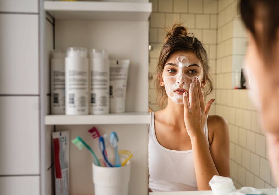 Young woman washing her face with a foaming cleanser while looking in a bathroom mirror