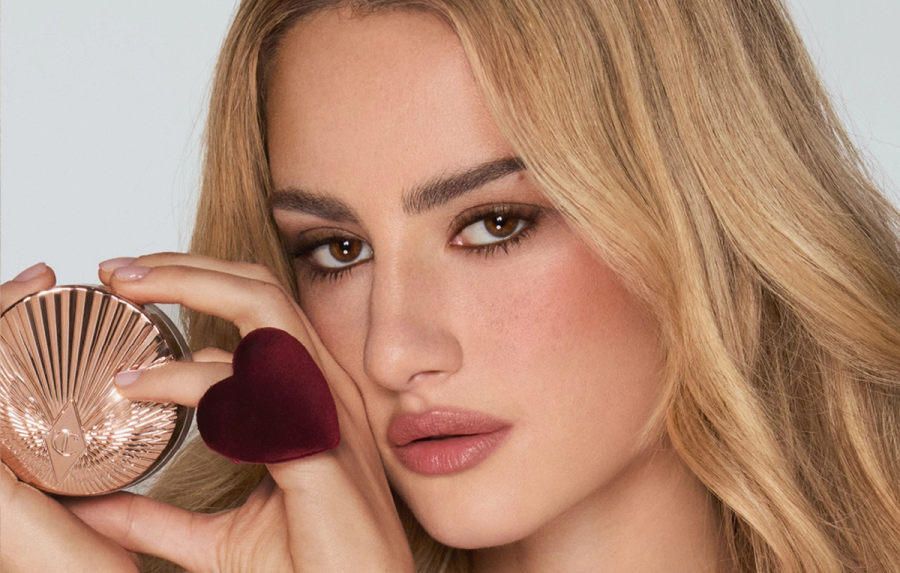 Grace Van Patten for Charlotte Tilbury, holding a shimmering rose-gold highlighter compact and a heart-shaped velvet applicator up to her cheek, with dewy, rosy makeup and nude-pink lips.