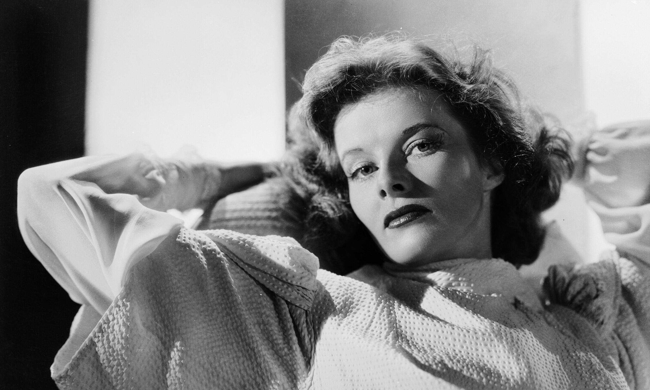 Black and white photo of Katharine Hepburn relaxes with her hands behind her head in 1938.