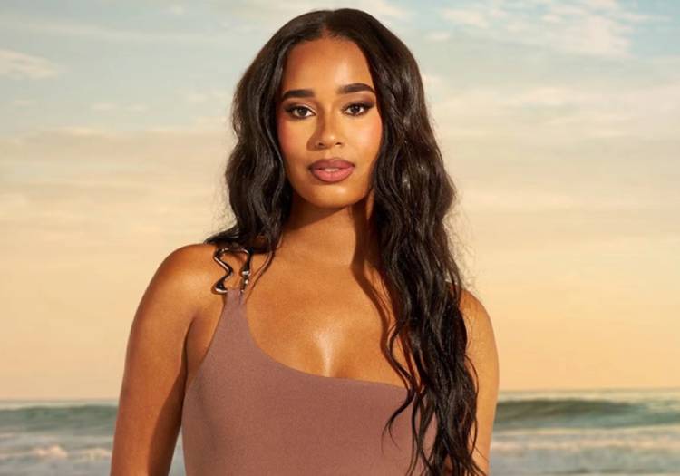  Summer House star Mia Calabrese poses on a beach at sunset wearing a taupe asymmetric one-shoulder swimsuit, with long dark wavy hair and a natural glowy makeup look
