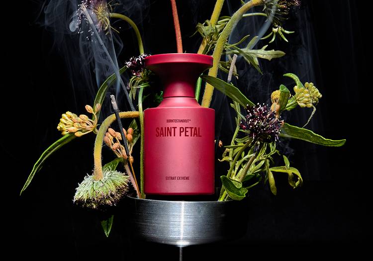 Red BORNTOSTANDOUT Saint Petal perfume bottle surrounded by flowers, green stems and smoking incense against a black background.