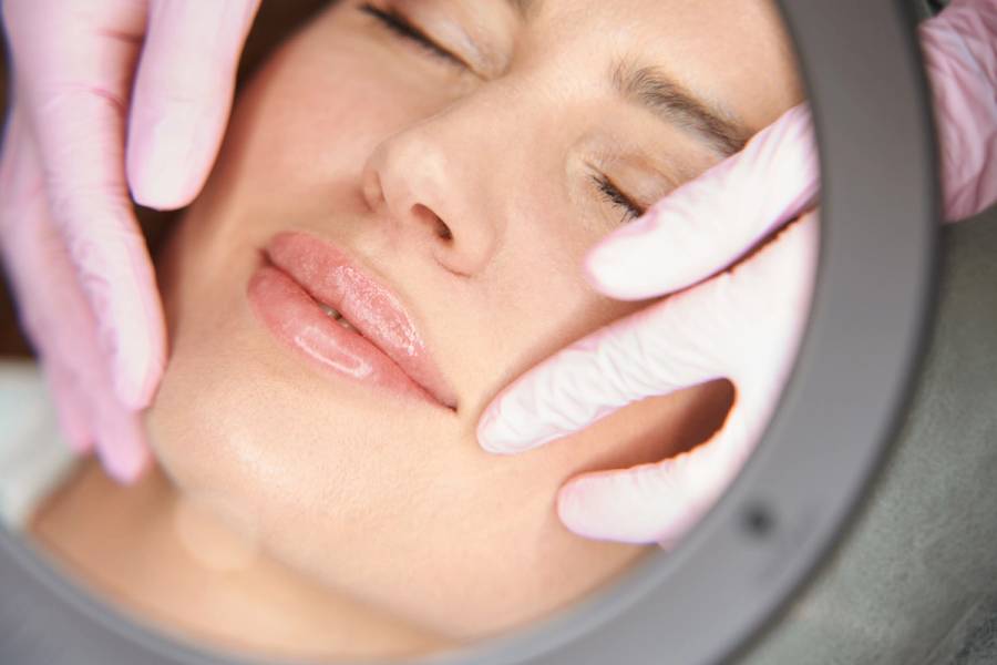 Close-up of a woman with her eyes closed as a provider in pink gloves holds her face during a lip treatment, seen through a lamp.