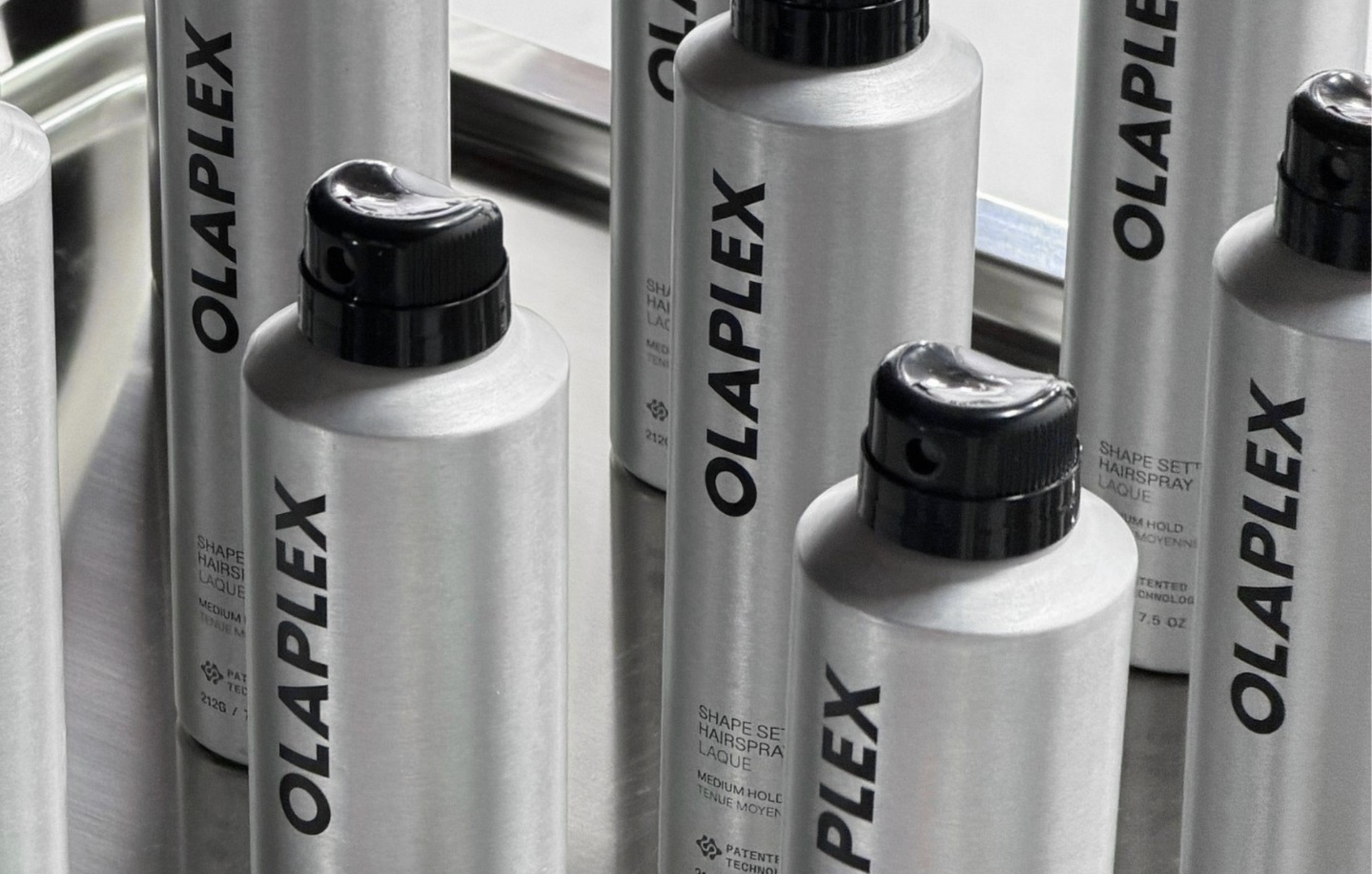 Several silver bottles of OLAPLEX Shape Set Hairspray arranged together in a black-and-white product shot.
